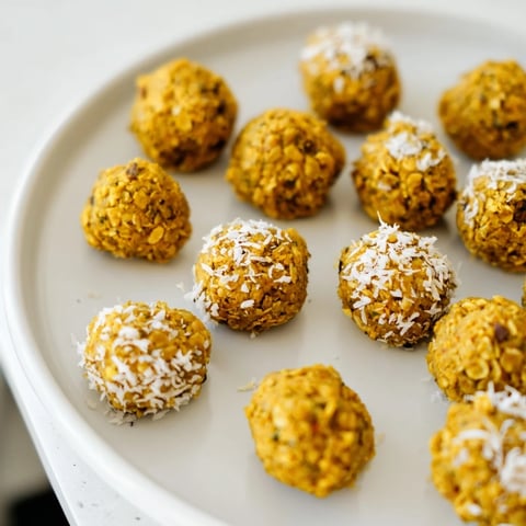 Freshly rolled Ginger Turmeric Energy Balls, coated in shredded coconut, display a vibrant orange hue from the turmeric and specks of grated ginger on a rustic wooden board.  